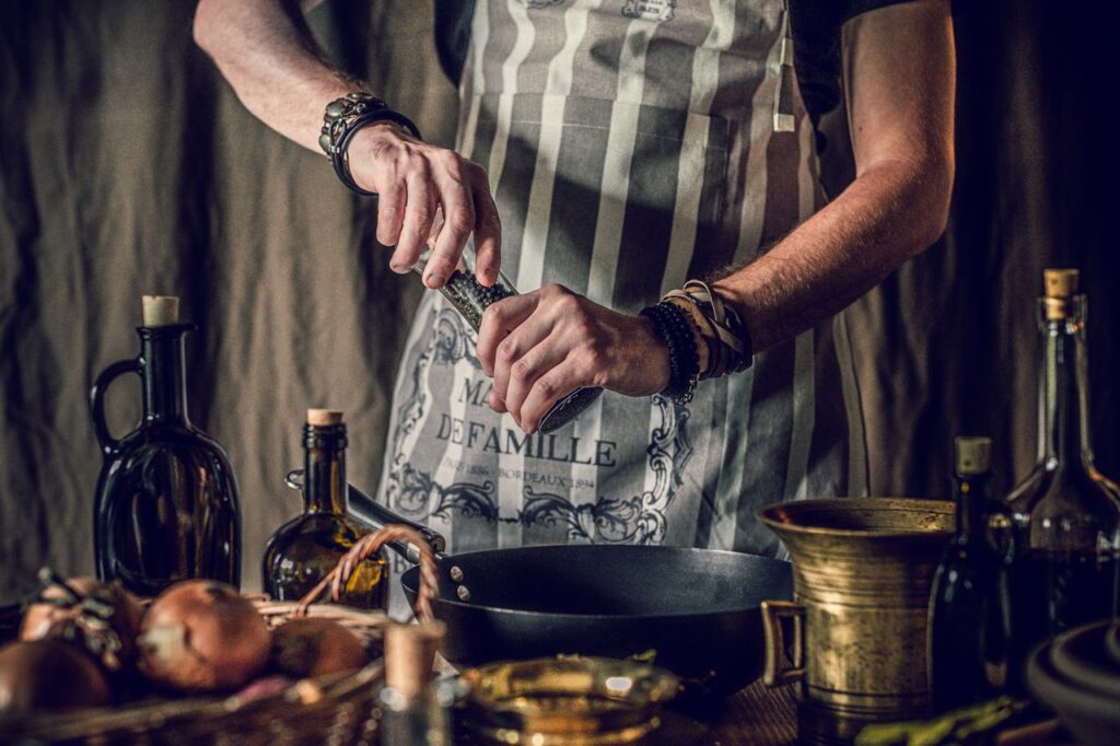 Crop faceless man in apron adding pepper in pan while cooking traditional dish standing at table with assorted ingredient and oil bottles in rustic kitchen